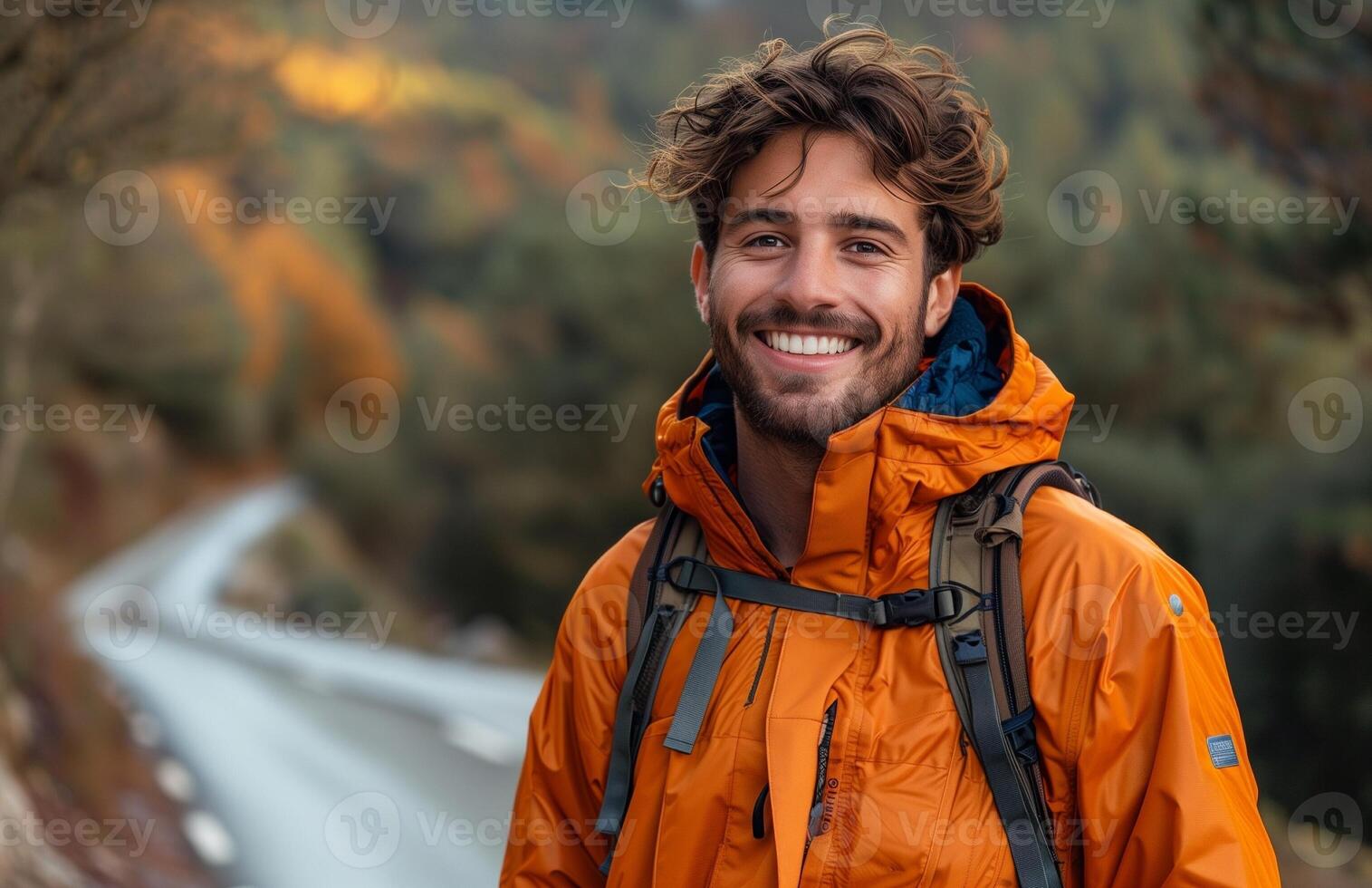 A cheerful man in an orange jacket stands on a road amidst lush greenery photo