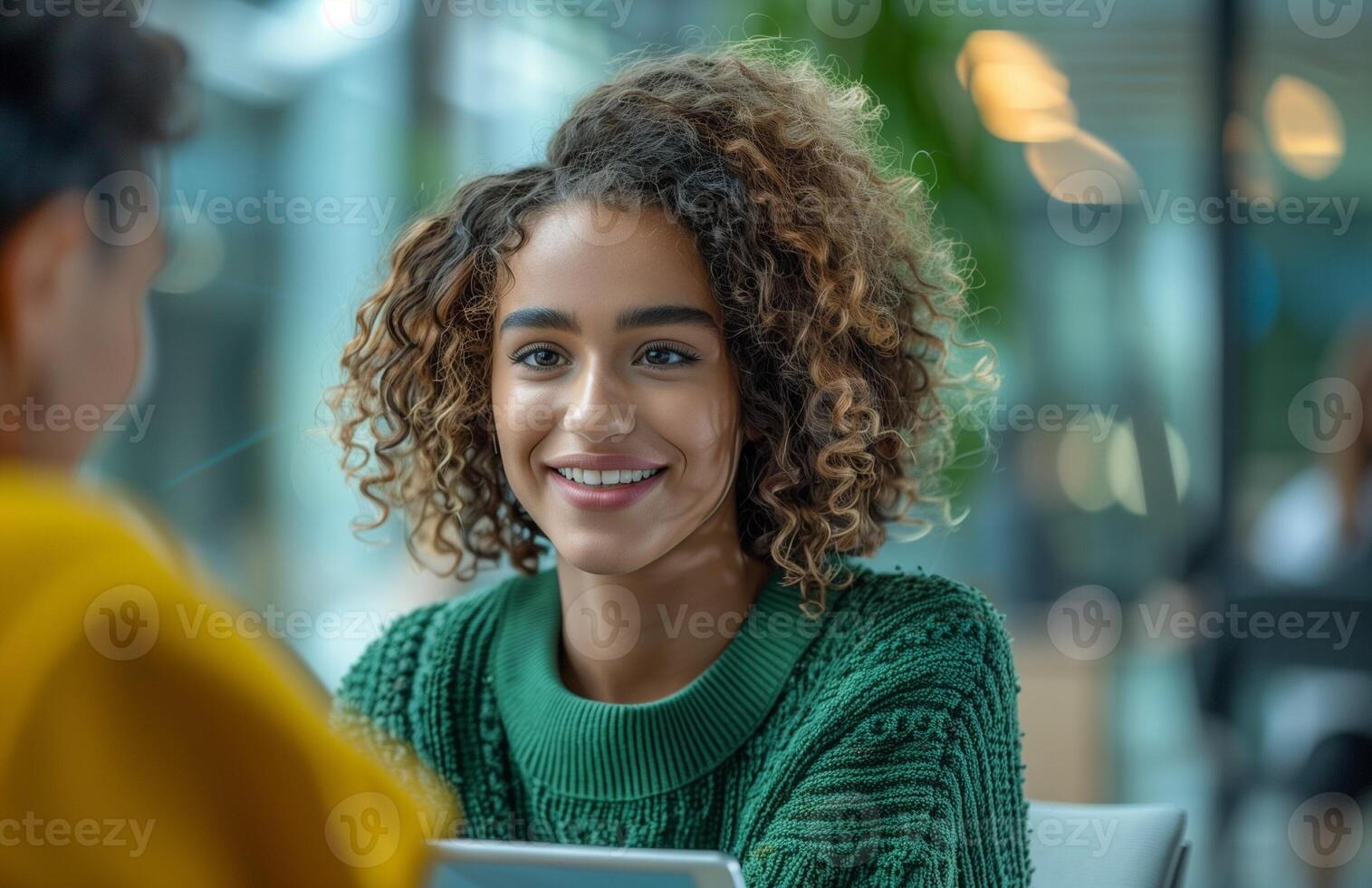 A curly haired young woman in a green sweater interviews in an office with a tablet photo