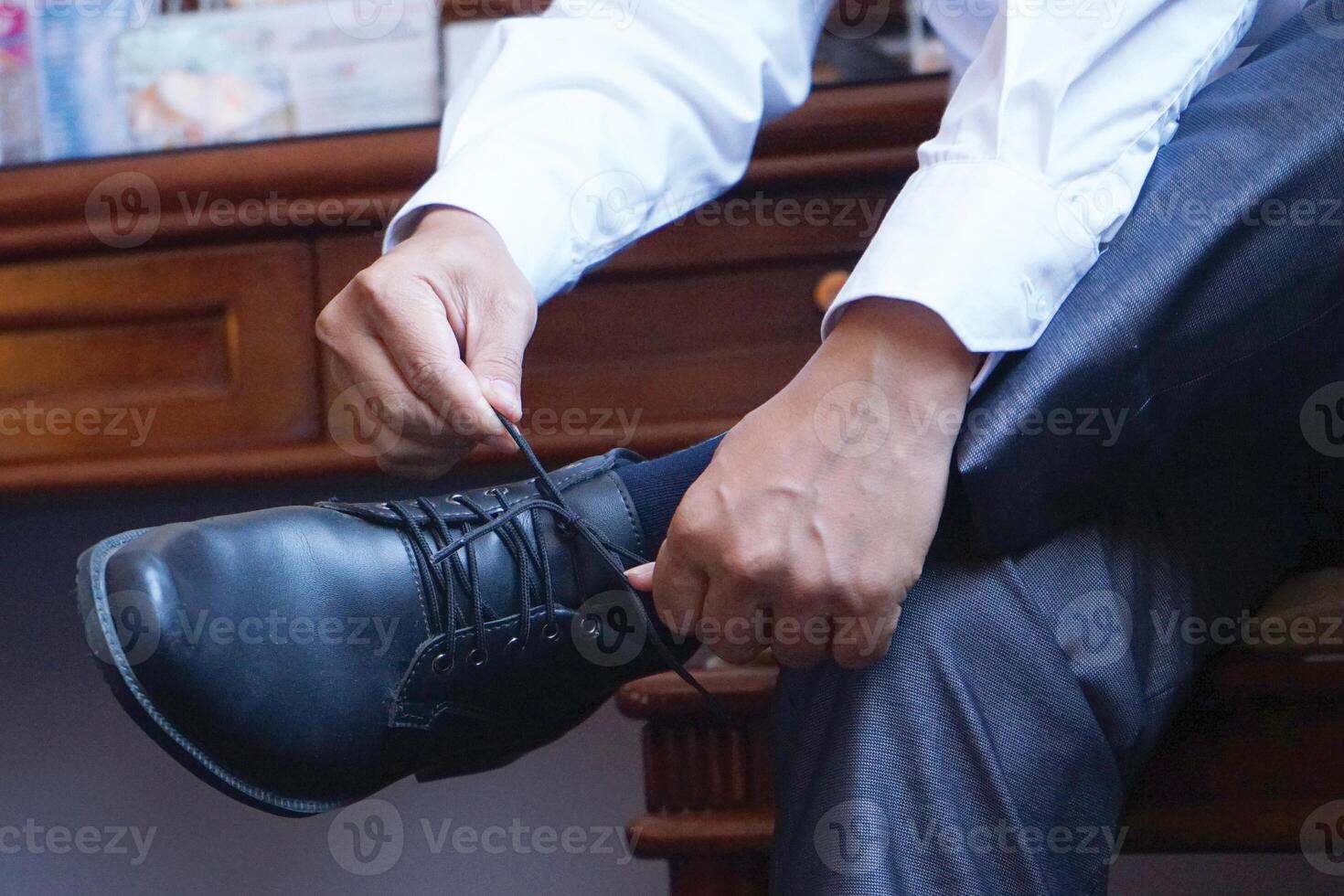 A man, the groom puts leather stylish shiny black shoes on his feet in socks in the morning. Sitting on the sofa and tying his shoelaces with his hands. Wedding photography, businessman portrait. photo