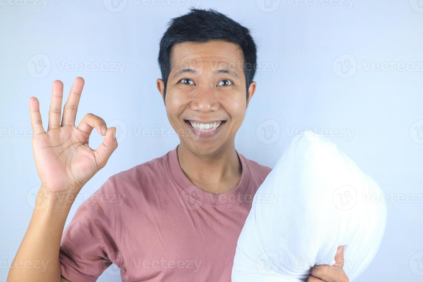 Smiling facial expression of young Asian man hugging pillow showing okay gesture. bedtime concept photo