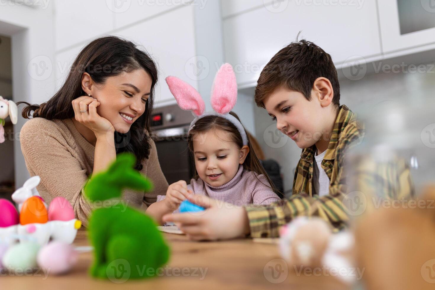 Cute little children wearing funny bunny ears headbands embracing and kissing young happy mother while painting Easter eggs together, sitting at table in kitchen, selective focus photo