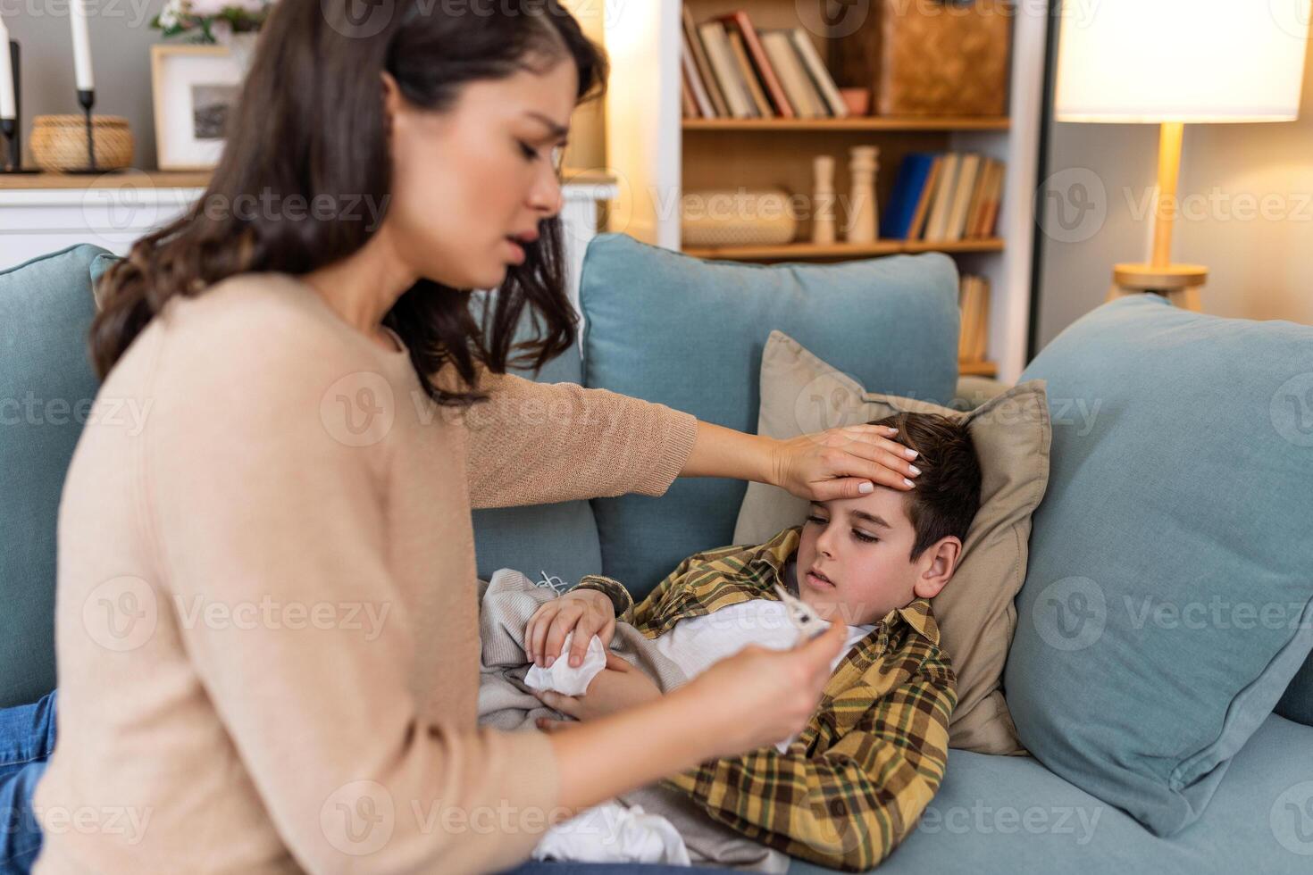 Young woman checking temperature with hand of her ill son. Mother checking temperature of her sick boy. Sick child lying on bed under blanket with woman checking fever on forehead by hand. photo