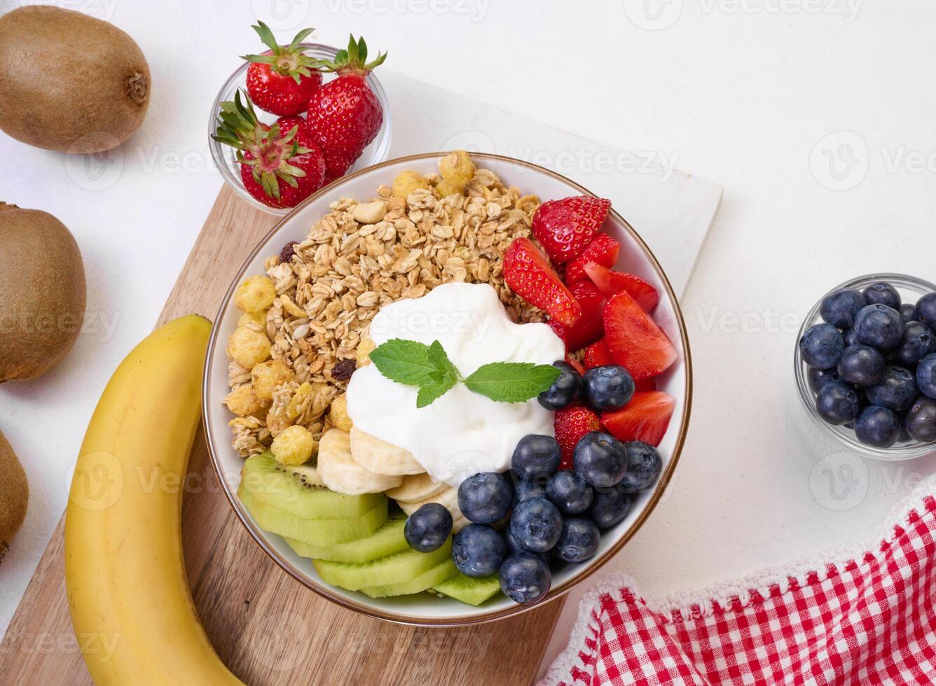 Granola with strawberries, kiwi, banana and blueberries in a round plate on a white table. photo