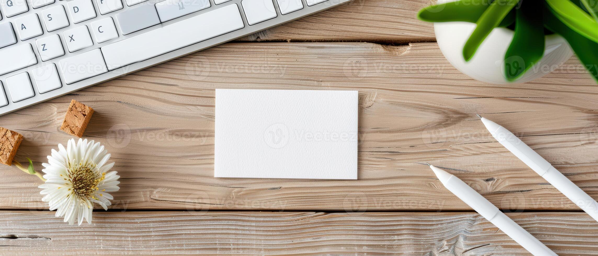 Top view of a clean desktop with blank white card, keyboard, pens, plant, and flower. Ideal for workspace and stationery themes. photo
