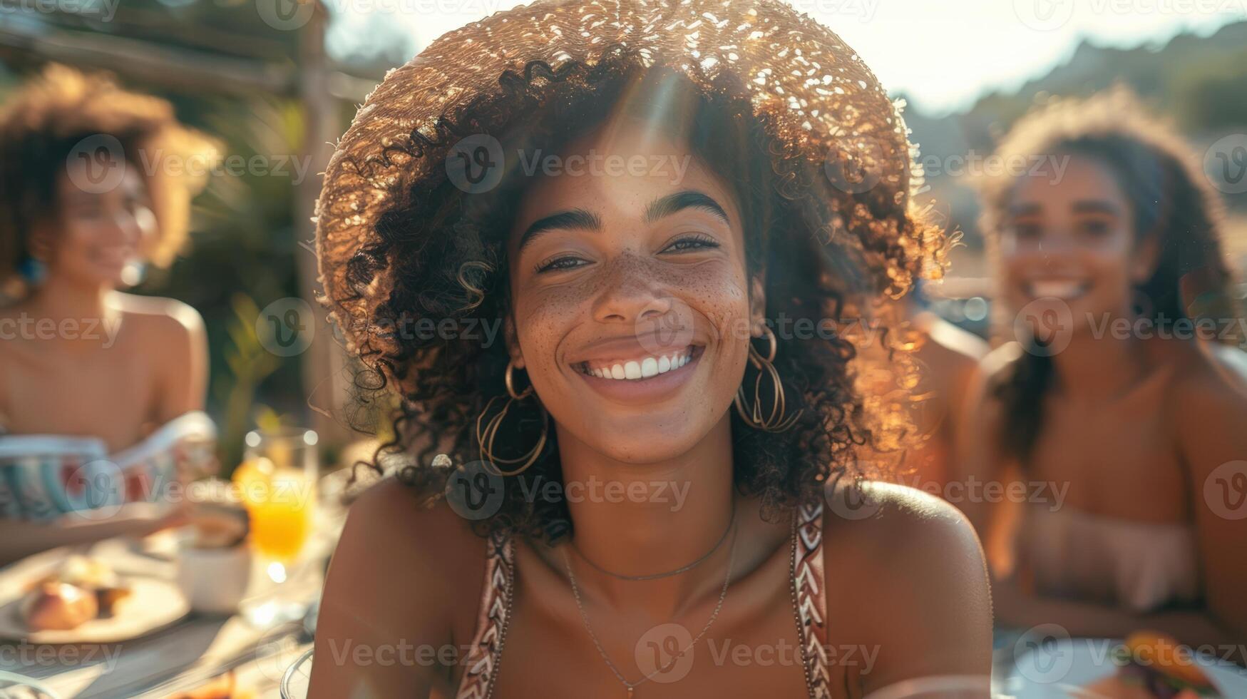A cheerful young woman with curly hair and a hat, smiling joyfully at an outdoor summer gathering with friends. photo