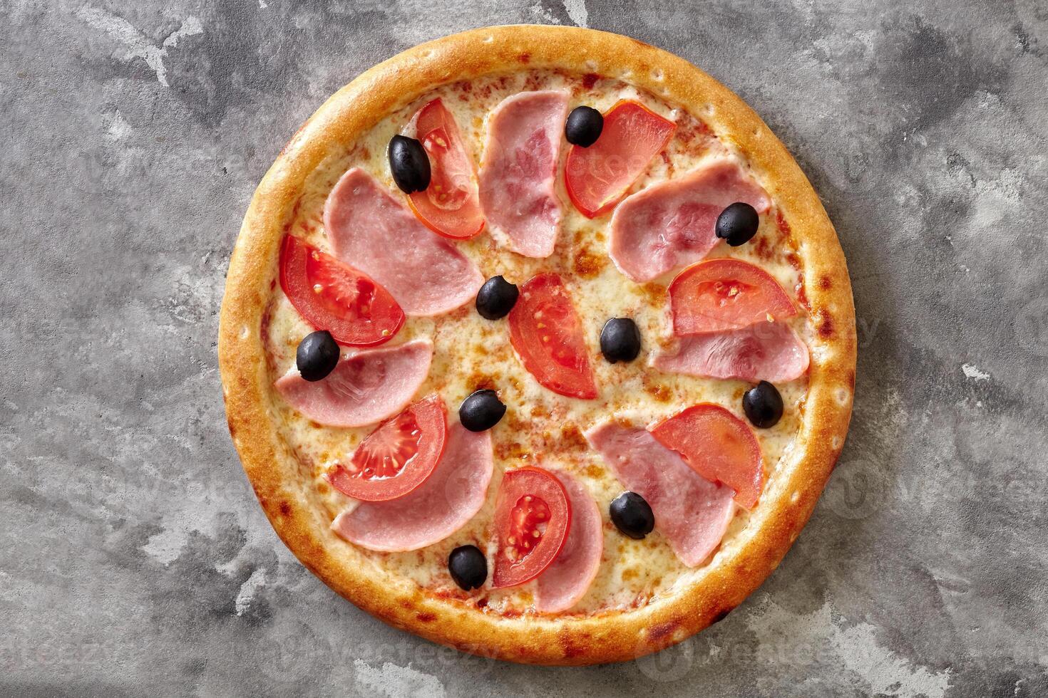 Thin classic pizza with melted cheese, ham, tomatoes and black olives on gray stone surface photo