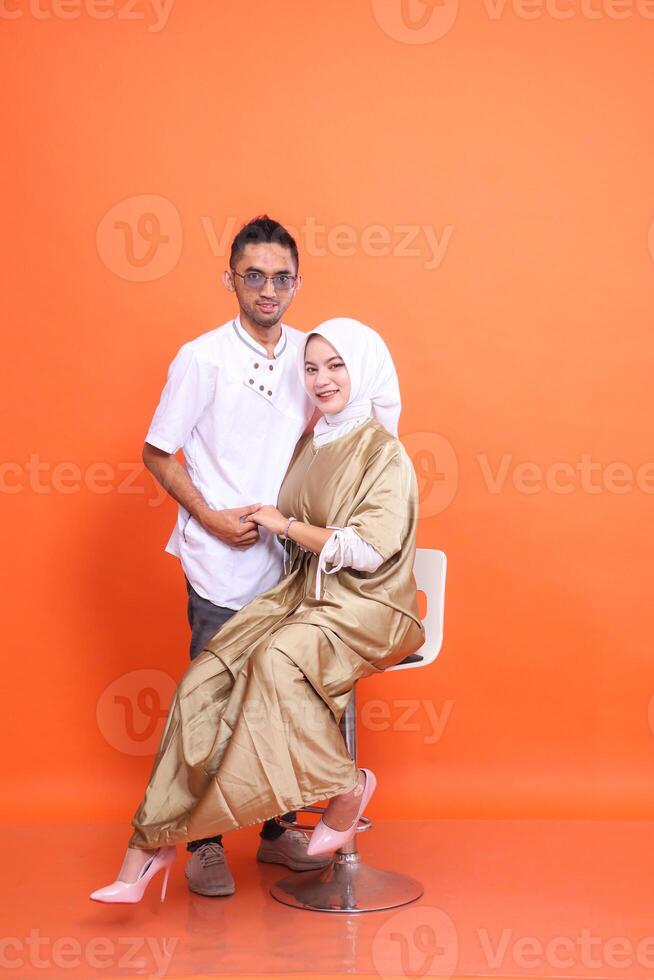 Asian muslim couple man and woman smiling holding hands wearing kaftans and muslim clothes in the month of Ramadan isolated orange background. Islamic religious, fashion, commercial concepts photo