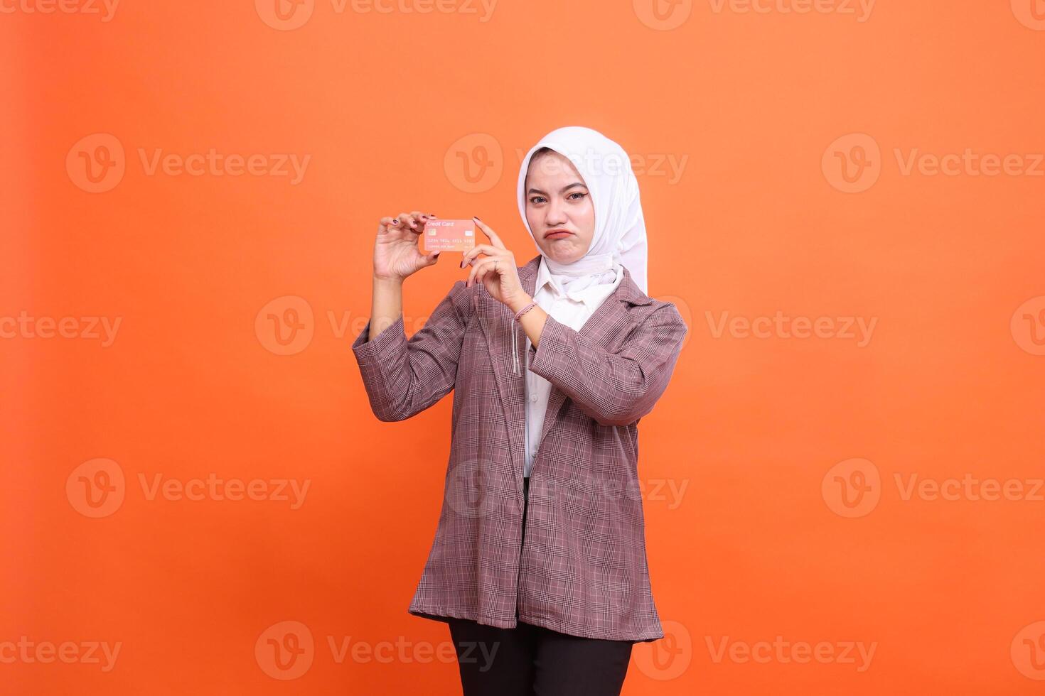 Young indonesia woman in hijab suit frowning both hands showing isolated plastic debit credit card on orange background. customer satisfied with service for financial , mobile transaction, payment photo