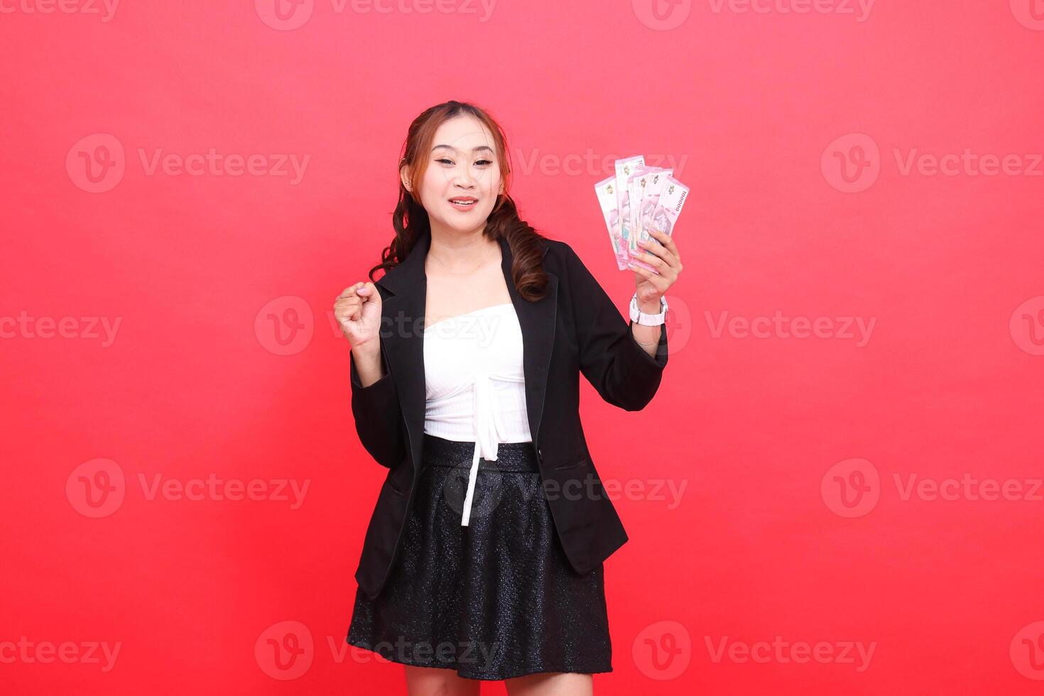 gesture of young Asian office female happily wearing black formal suit, hand holding rupiah money while clenching it, for financial content, business, advertising, transactions photo