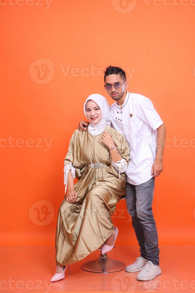 indonesia muslim couple man and woman smiling embracing on shoulders wearing kaftans and muslim clothes in the month of Ramadan isolated orange background. Islamic religious, fashion, advertising photo