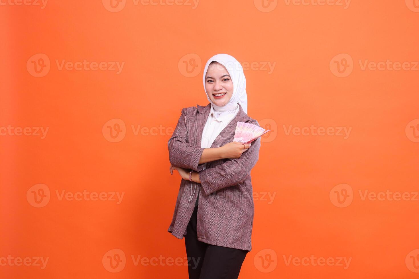 Charm of beautiful Asian woman wearing cheerful hijab with arms crossed holding rupiah banknotes to the side isolated on orange background. finance, lifestyle, advertising concept photo