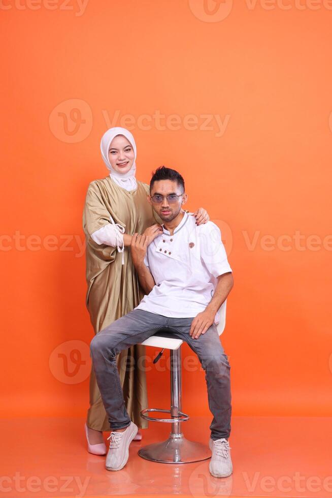 indonesia muslim couple man and woman smiling holding his man's shoulders wearing kaftan and muslim clothes in the month of Ramadan isolated orange background. Islamic religious, advertising photo