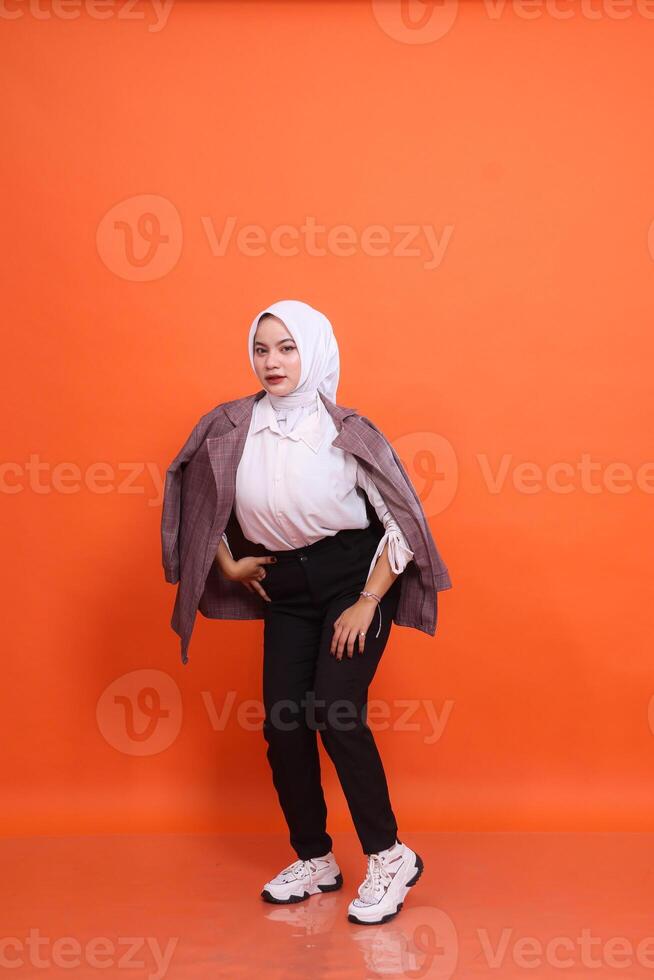 Full length portrait of beautiful elegant Muslim indonesia woman model holding pockets and thighs wearing office clothes wrapped in jacket isolated orange studio background. Concept of fashion, beauty photo