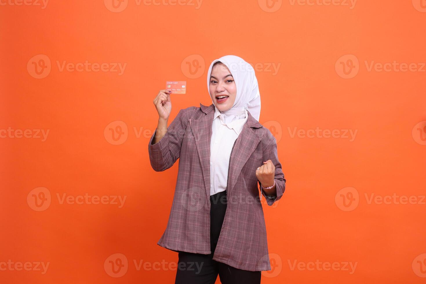 mature indonesia woman hijab cheerful suit holding plastic debit credit card while clasping hands isolated on orange background. satisfied with service for financial concept, transactions, payments photo