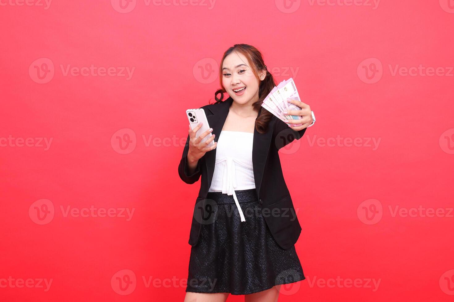 expression of young asian career cheerful woman wearing black formal suit, hand holding rupiah money and mobile phone in front, for technology, business, advertising, financial content photo