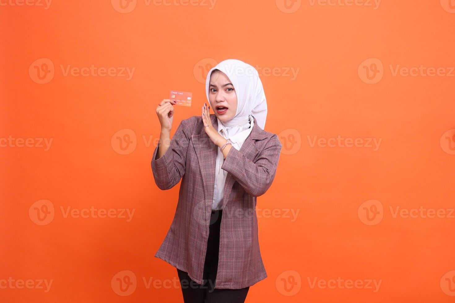 mature asian woman in hijab suit shocked holding plastic debit credit card while hands whispering isolated on orange background. satisfied with service for financial concept, transaction, service photo