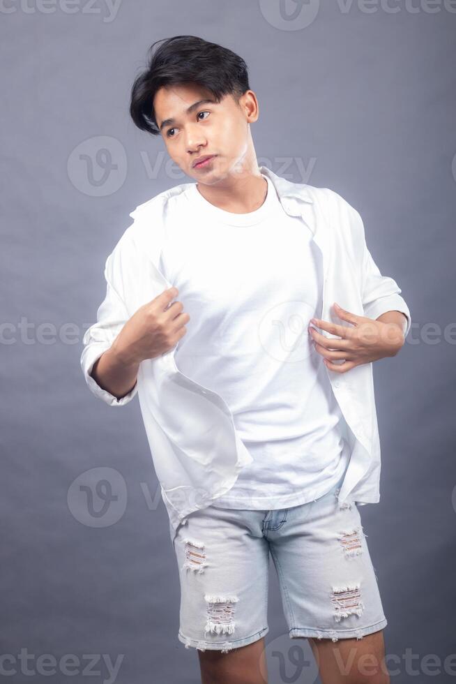 cool asian man posing standing head tilted left forward in front of the camera while holding his shirt in both hands indoors. fashion promotion man shirt photo session in the gray background