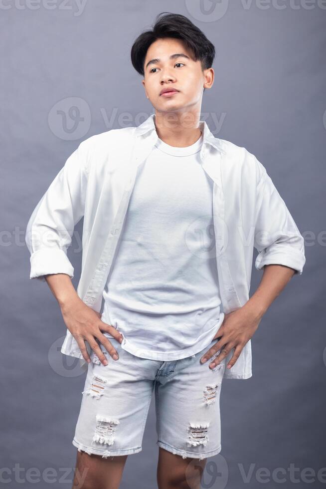 elegant asian man posing standing head to right in front of the camera while his ladder holding his pants indoors. fashion promotion man shirt photo session in the gray background studio