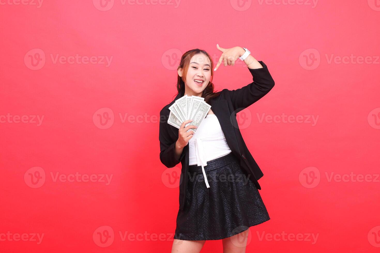 gesture of cheerful young asian office girl wearing black formal suit holding dollar bill and pointing at it from above, for corporate content, fashion, advertising, broadcast photo