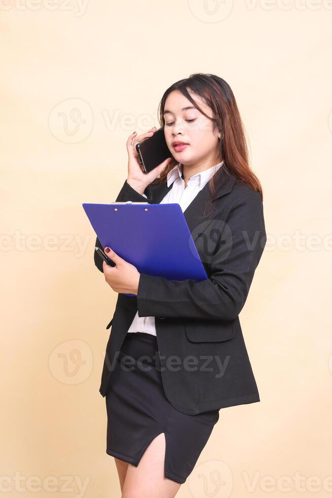 Beautiful formal Asian executive woman in suit standing and posing holding clipboard and telephone with gadget smartphone on yellow background for commercial, vacancy and business photo
