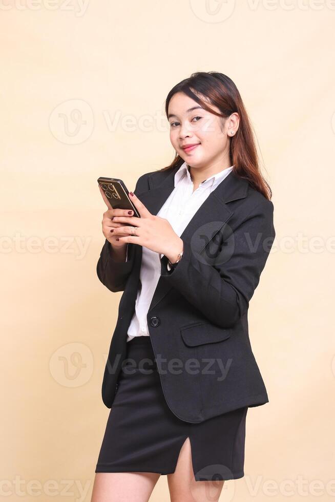Beautiful formal Asian executive woman in suit standing smile and holding while operating online shopping gadget smartphone on yellow background for commercial, vacancy and business photo