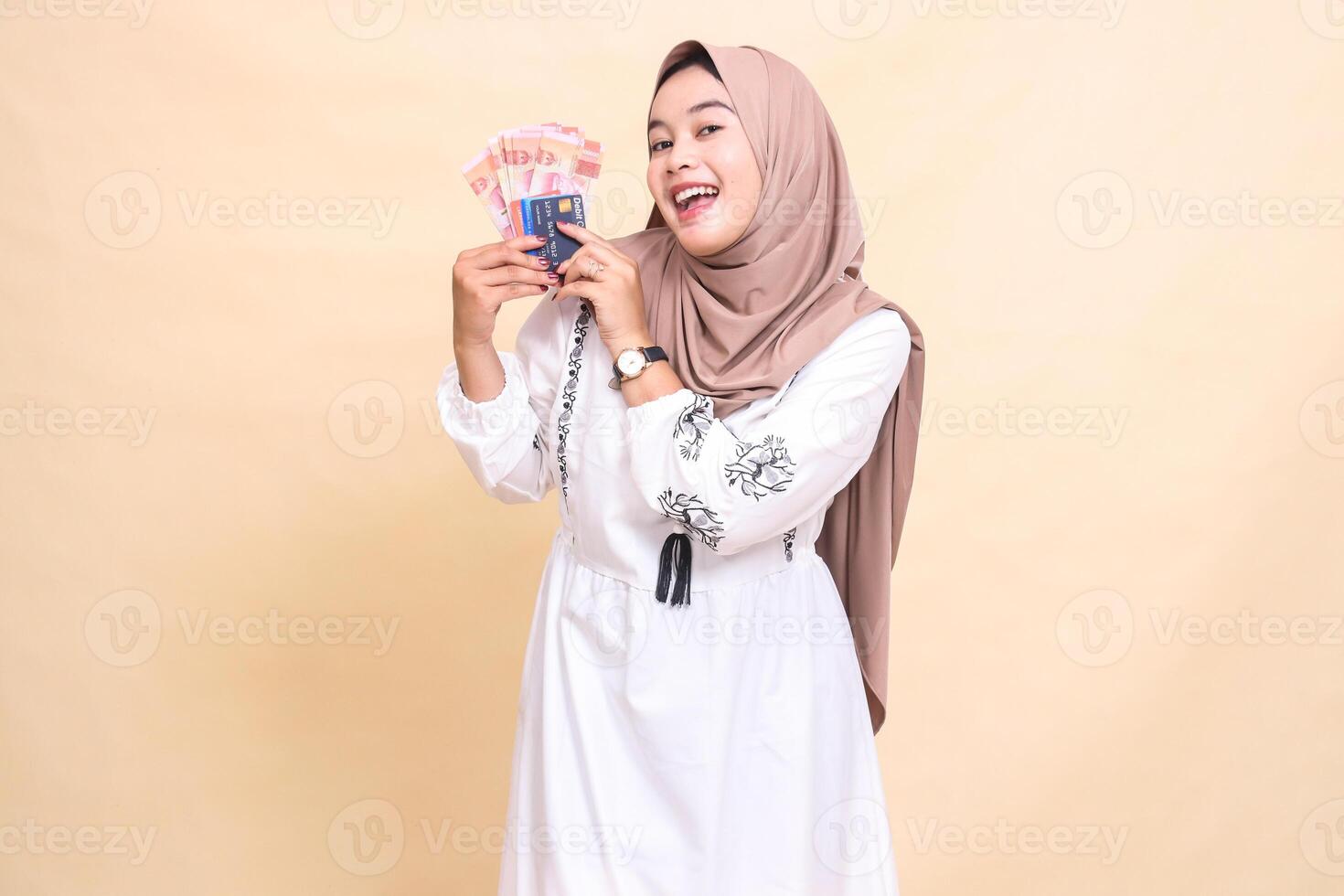 a beautiful indonesia Muslim woman wearing a hijab with a cheerful smile brings to her right side rupiah notes and a debit credit card for payment. used for advertising, technology, Eid and Ramadan photo