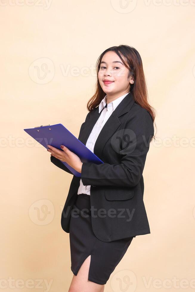 Beautiful formal indonesia executive mature girl in suit elegant smile standing and pose holding clipboard to camera, 3 by 4 body portrait on yellow background for commercial, vacancy and business photo
