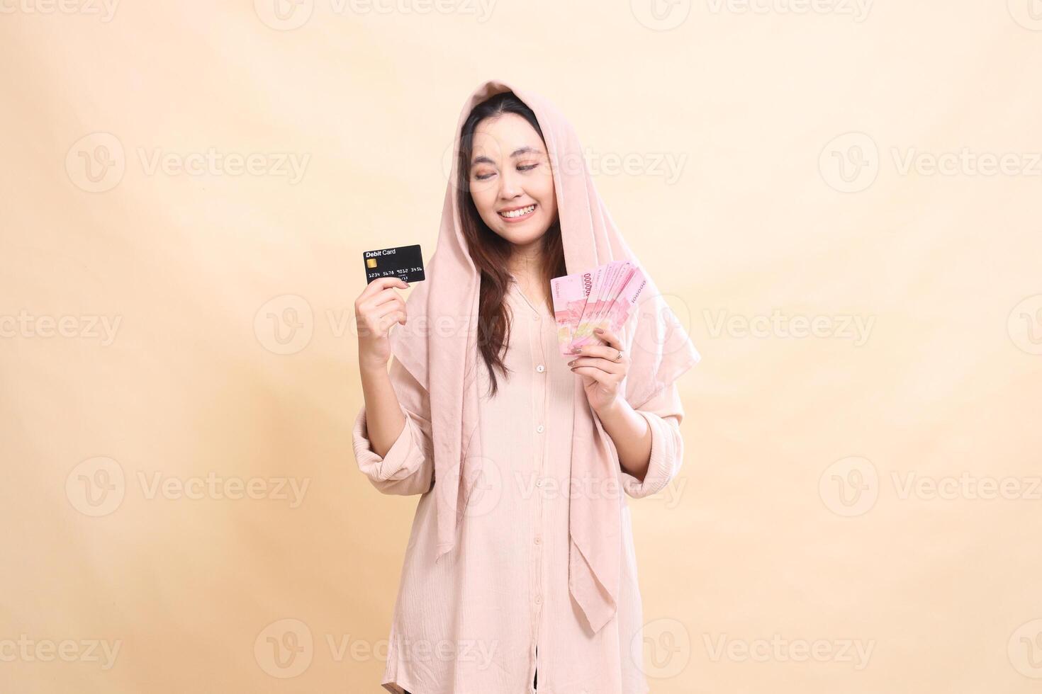 girl Asian with candid, graceful smile expression, hand holding debit card up and carrying rupiah money. young beautiful for finance, advertising, fashion photo