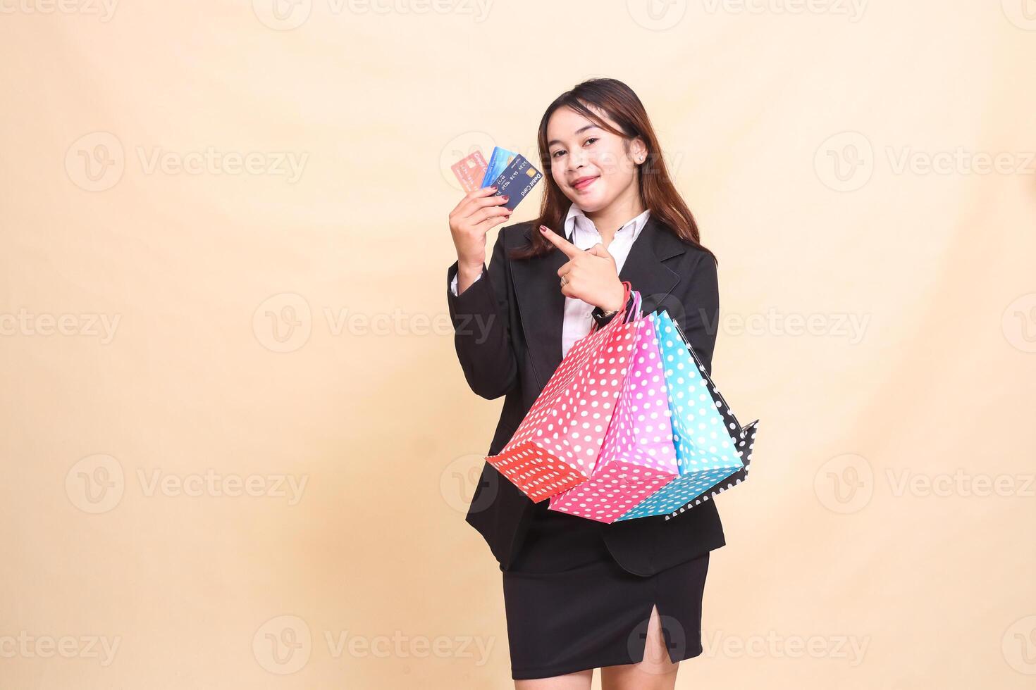 Beautiful formal Asian office woman in suit standing happy and carrying paper bag while pointing at debit credit card to start paying online on yellow background for commercial and business photo