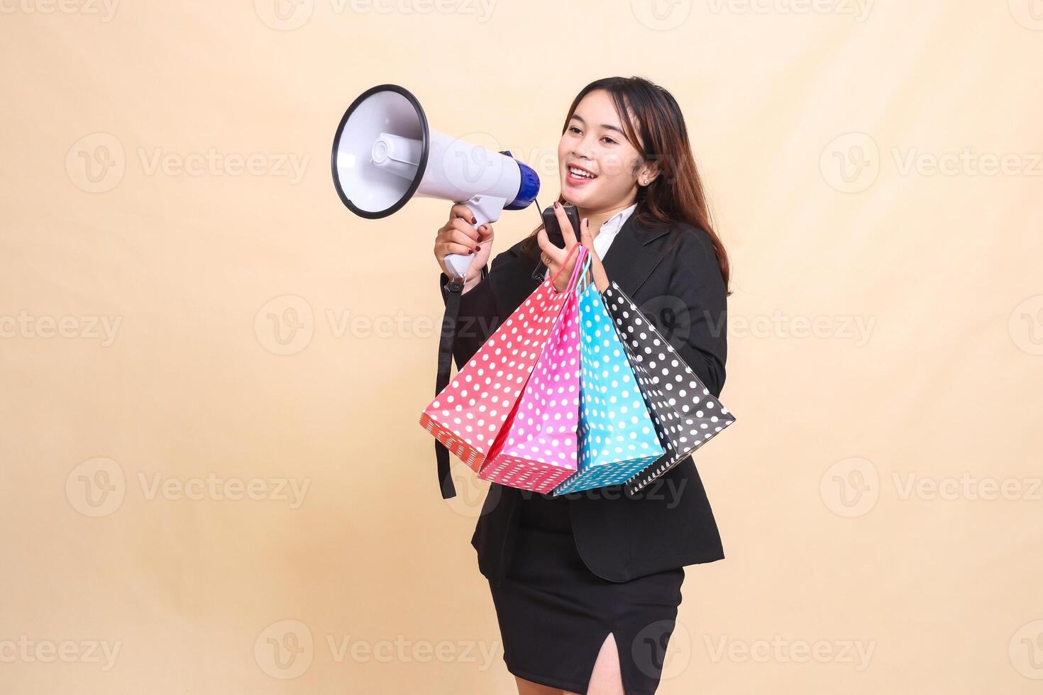 Beautiful formal Asian office woman in suit standing smiling happily carrying shopping paperbag while carrying mic using megaphone for promo announcement on yellow background for advertising photo