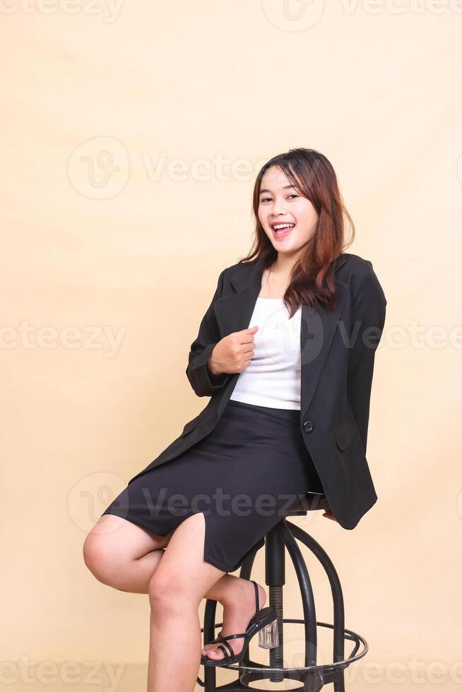 Beautiful cheerful young Asian executive woman sitting on chair and pose holding jacket and chair to camera, 3 by 4 body portrait on yellow background for commercial, technology and business photo