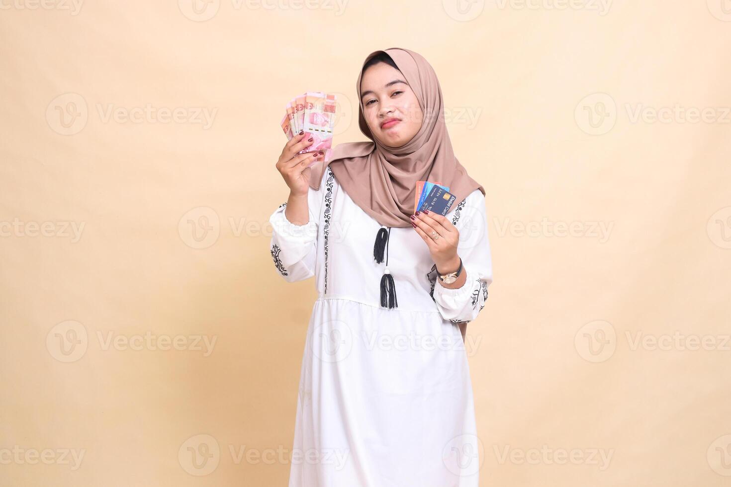 a beautiful Asian Muslim woman wearing a sad hijab frowns carrying rupiah money and a debit credit card for payment. used for advertising, technology, Eid and Ramadan photo