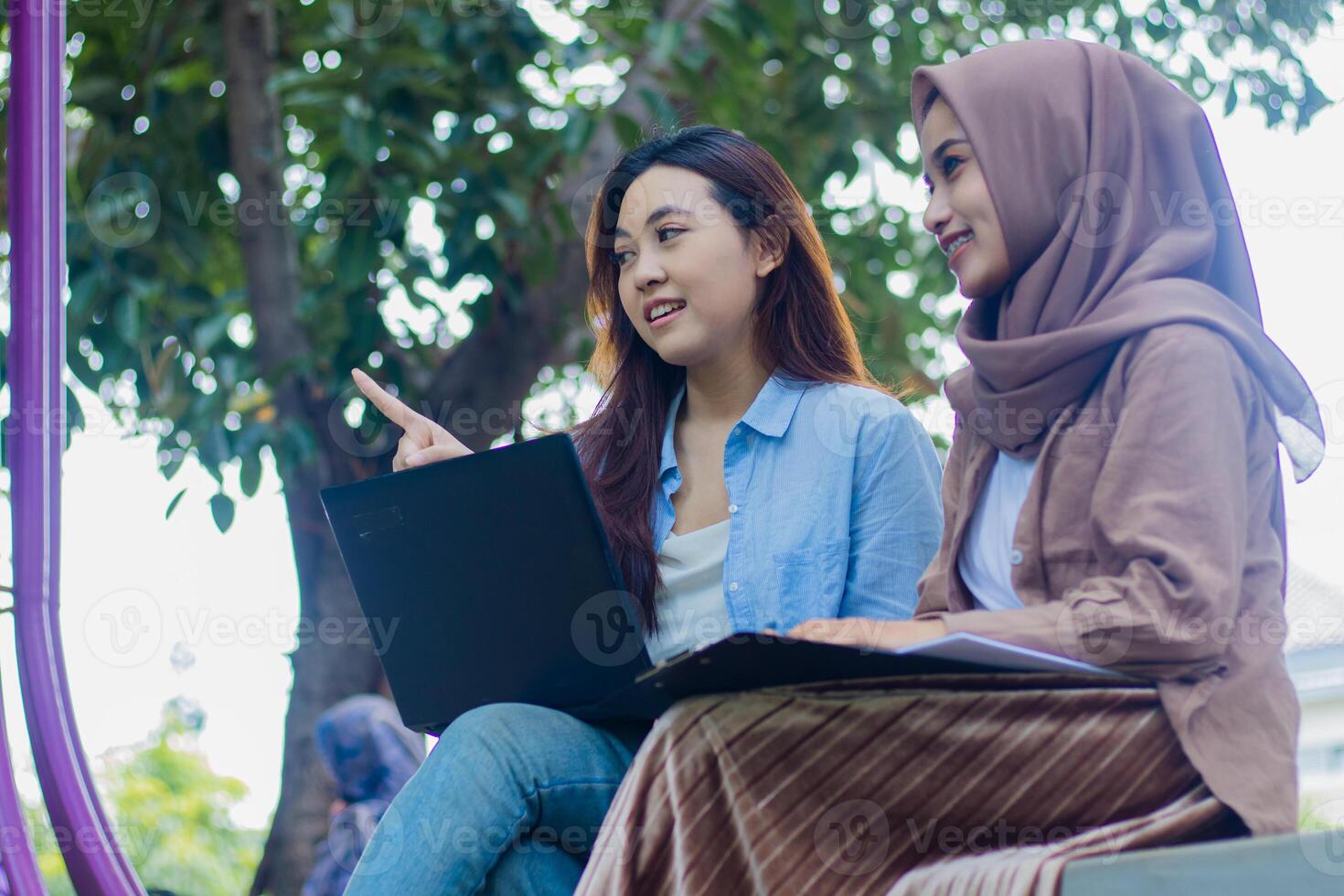 A pair of beautiful Asian women candidly show something left and right while chatting with a laptop, books and clipboard in the campus park. female students for education and lifestyle concepts photo