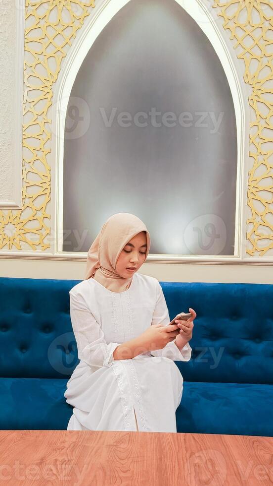 beautiful Asian woman wearing a hijab candid elegant expression checking her cellphone gadget sitting on the sofa with a background of calligraphy ornaments of an Islamic mosque in Arabic nuances photo