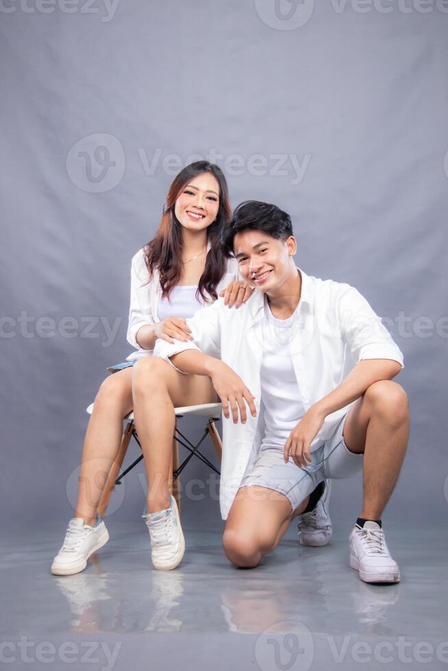 smiling man and woman posing sitting in front of the camera while their ladders holding their thighs facing forward indoors. fashion promotion couple photo session in gray background studio