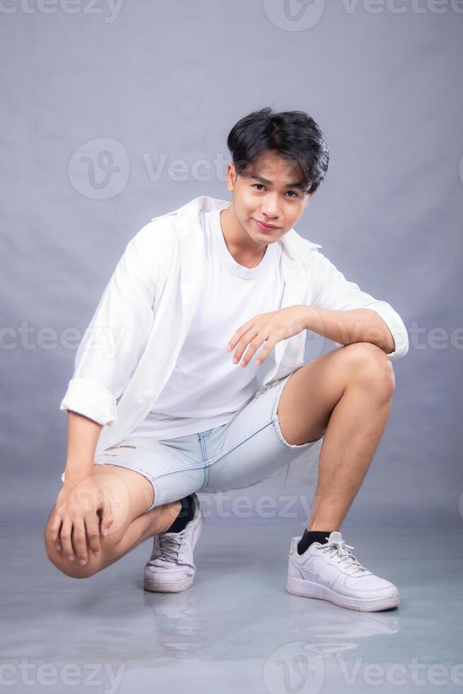 elegant asian man indonesian is posing crouching sideways in front of the camera while his hands are on his knees indoors. fashion promotion man shirt photo session in the gray background studio