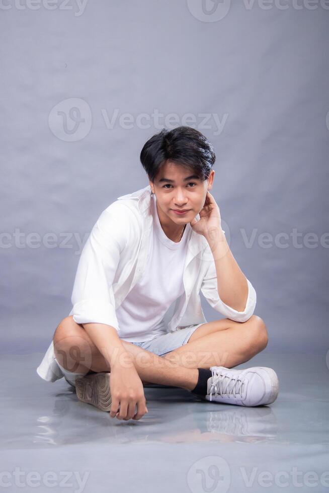 elegant asian man posing sitting in front of the camera while holding his left cheek indoors. fashion promotion man shirt photo session in the gray background studio