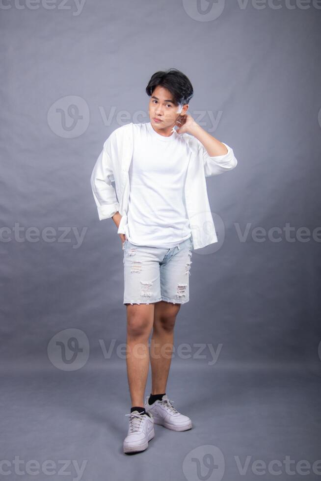 elegant asian man posing standing head tilted in front of camera while hands back indoors. fashion promotion man shirt photo session in the gray background studio