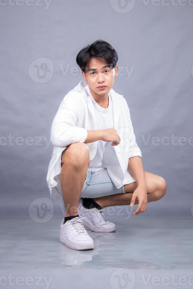 elegant asian man posing squatting upright in front of the camera while his hands are on his knees indoors. fashion promotion man shirt photo session in the gray background studio