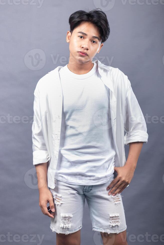 elegant asian man posing standing with his head straight forward in front of the camera while holding his pants indoors with both hands. fashion promotion man shirt photo session in the gray