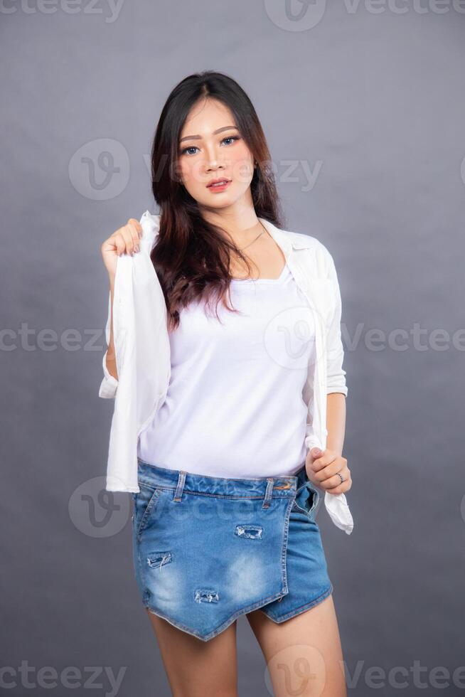 elegant asian woman posing standing in front of the camera while holding her shirt in her hand. fashion promotion woman shirt photo session in gray background studio
