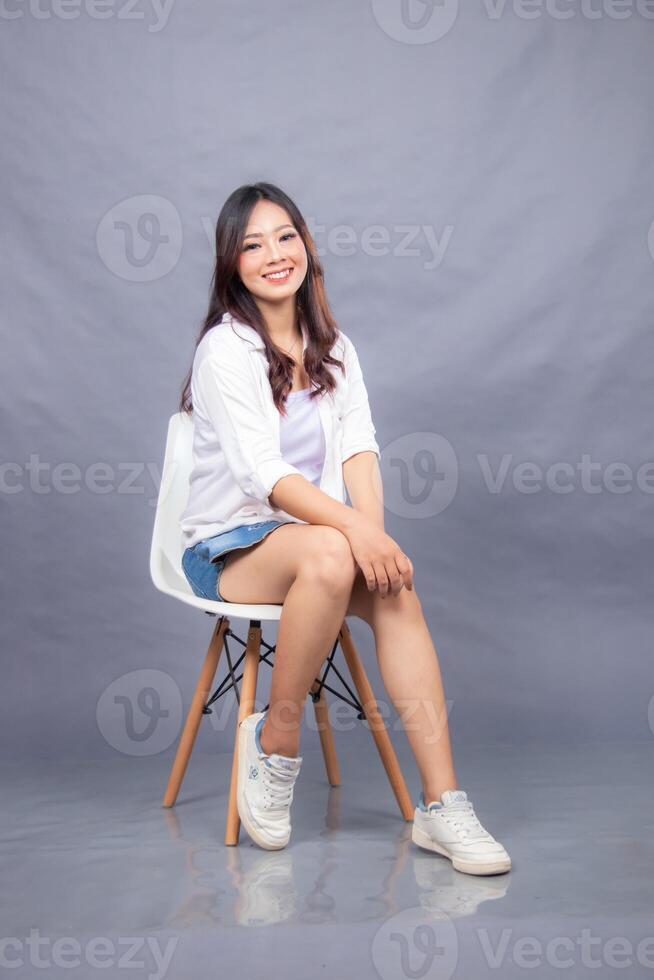 cheerful smiling woman indonesian posing sitting on a chair in front of the camera while her ladder leans forward indoors. fashion promotion woman shirt photo session in gray background studio