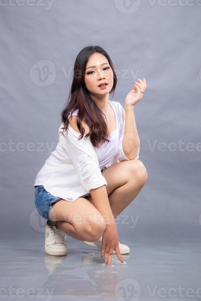 elegant woman is posing squatting in front of the camera while her ladder is holding on to the floor indoors. fashion promotion couple shirt photo session in gray background studio