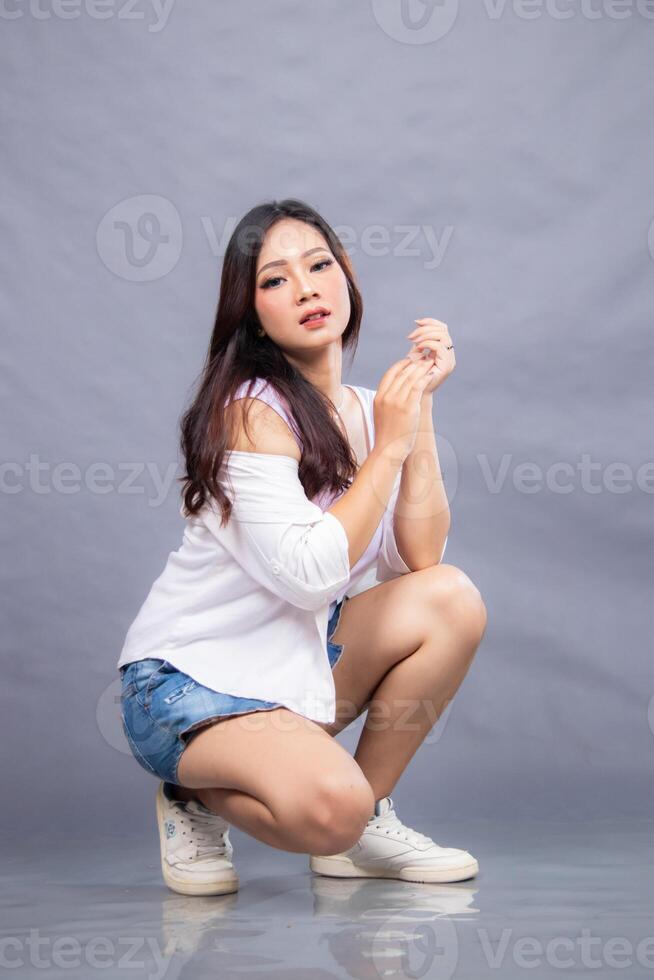 an elegant woman is posing squatting in front of the camera while her ladder is holding on indoors. fashion promotion couple shirt photo session in gray background studio