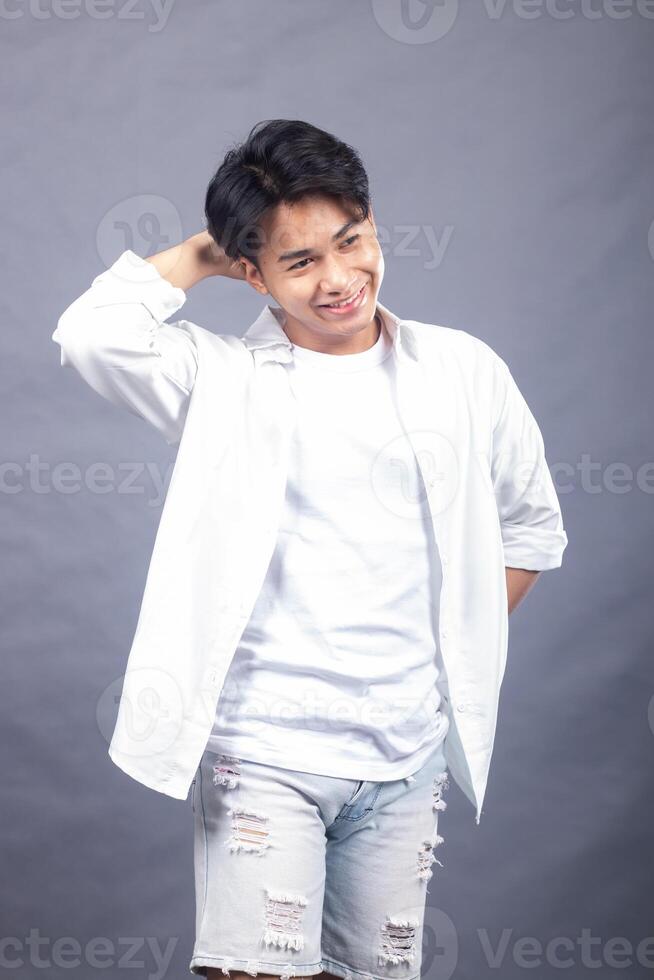 elegant smiling asian man posing standing with his head held high in front of the camera while holding his hair in a ladder indoors. fashion promotion man shirt photo session in the gray background