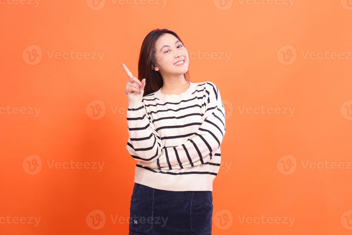 Portrait of a beautiful cheerful confident asian girl standing with arms crossed pointing top right and looking at the camera isolated on orange background wearing white sweater photo
