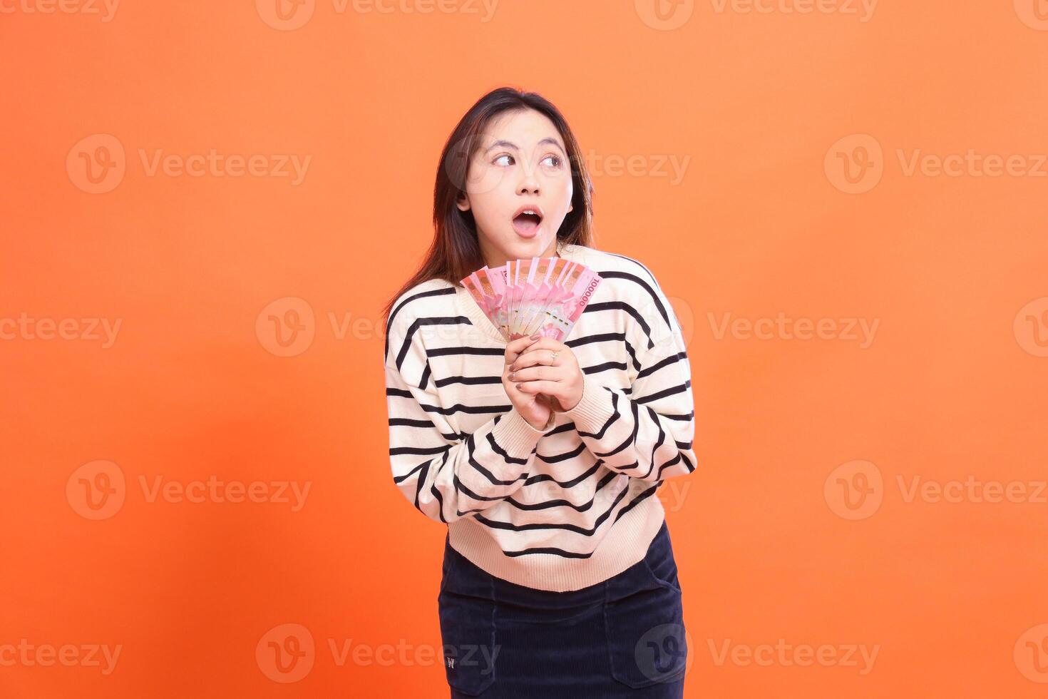 an Asian woman with a candid shocked expression standing while both hands holding several rupiah bills. for fashion, business and finance concept photo