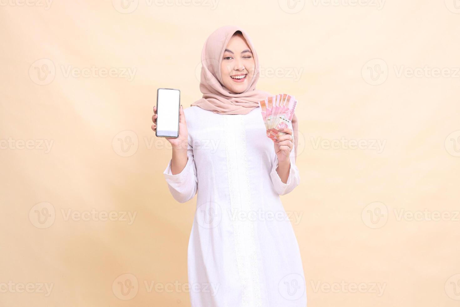 woman asian muslim wearing a hijab, smiling cheerfully, winking, holding a cellphone, gadget and rupiah money in her hand. Lifestyle, business and commercial concept photo