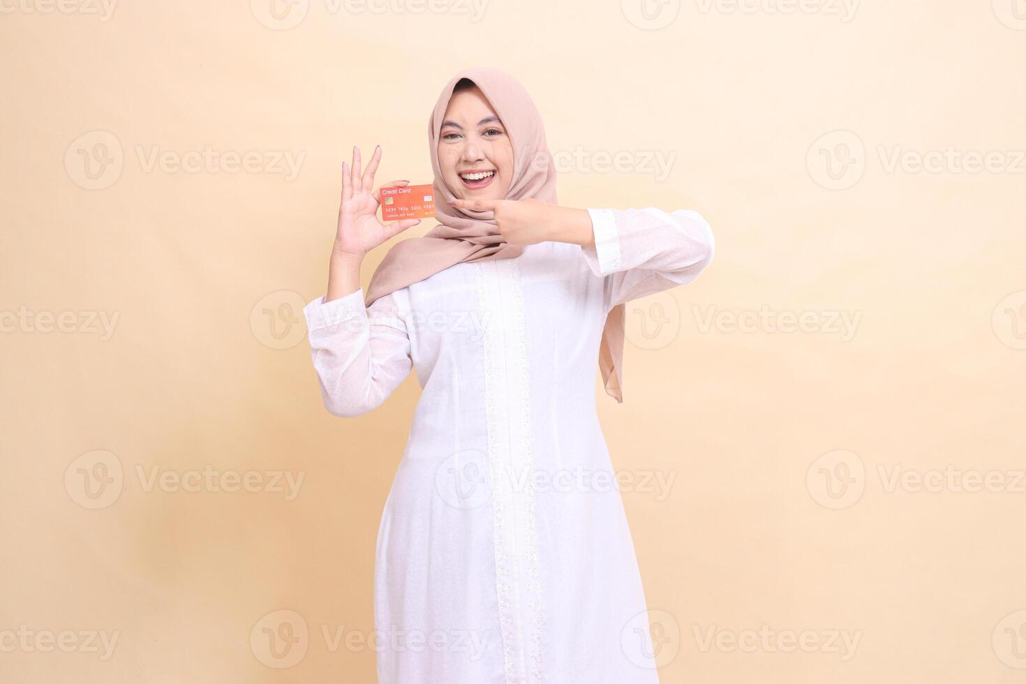 woman indonesia mature wearing a hijab smiles cheerfully holding a debit credit card in her hand and pointing up. Lifestyle, business and finance concept photo