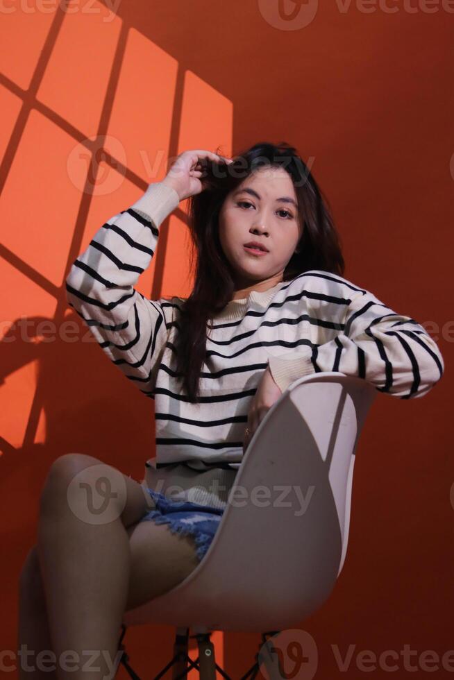 A beautiful elegant asian girl holds her hair sitting on a chair in a dark studio, the light shines on her forming a window pattern. Portrait of woman in projection light spotlight in studio photo